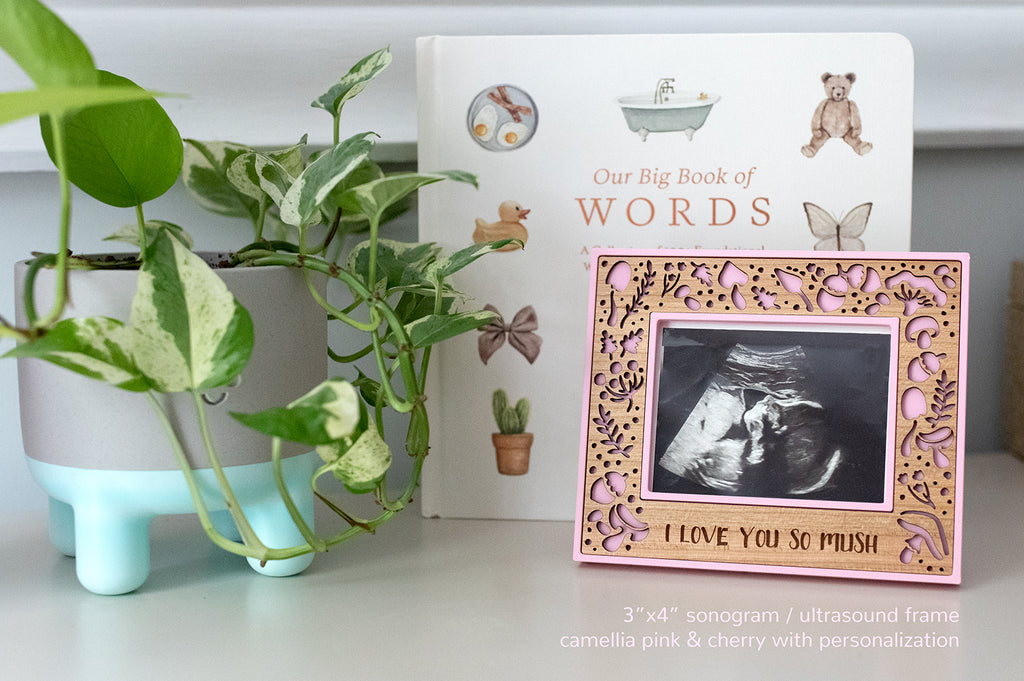 A small ultrasound picture frame displaying a photograph of a new baby sonogram image. The frame is pink with a unique mushroom design. There is a baby book and greenery sitting by the frame.