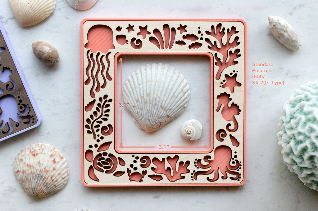 A standard polaroid beach frame with cute laser cut sea creatures. The frame opening is 3.1" x 3.1" and is surrounded by seashells and a blue coral candle. The frame is magnetic and has a pink coral backdrop.