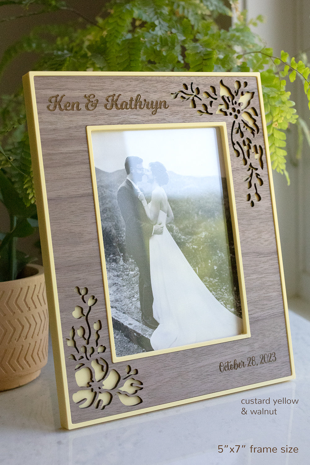A 5x7 picture frame in vertical orientation and made with pale yellow 3D printed PLA and an intricate laser cut walnut wooden inlay. The frame displays a photo of a newlywed couple embracing at their wedding.
