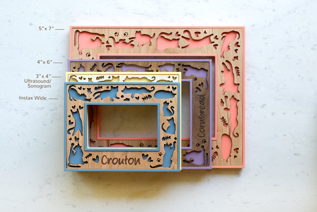 Cat photo frames with laser cut designs and engraved cat names. The frames are available in 5x7 in, 4x6 in, 3x4 in, and Instax Wide sizes. The top frame is slate blue with a cherry wood inlay and has a laser engraved cat name, "Crouton" on the bottom of the frame.