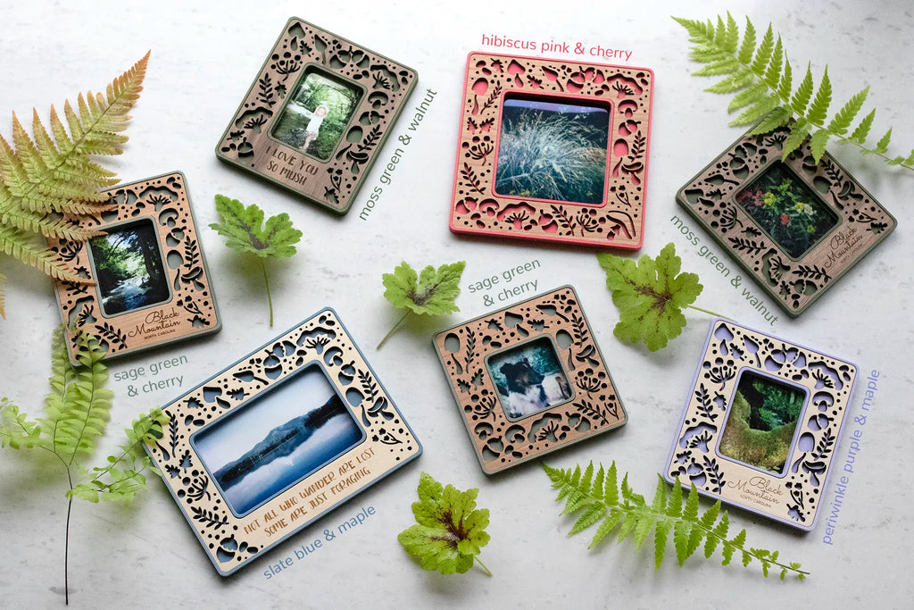 An assortment of colorful, laser cut mushroom magnetic photo frames. These polaroid frames are lying on a white surface, surrounded by greenery and ferns. The laser cut wooden inlay has an intricate, feminine design and is available in three wood types: walnut, cherry, and birch. Some of the frames include an engraved personalized message.