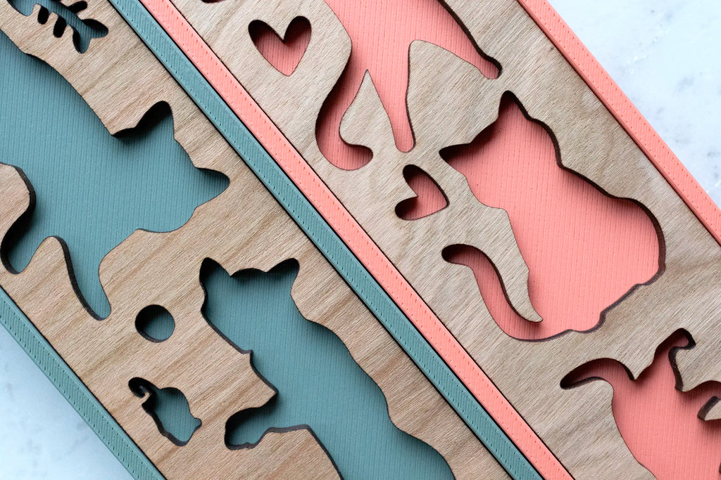 Detail of the sage green and coral cat picture frames up close, showing laser-cut cherry wood cat silhouettes and hearts layered over the textured 3D-printed backing of each handcrafted pet photo frame. Wood grain variation can be seen up close.