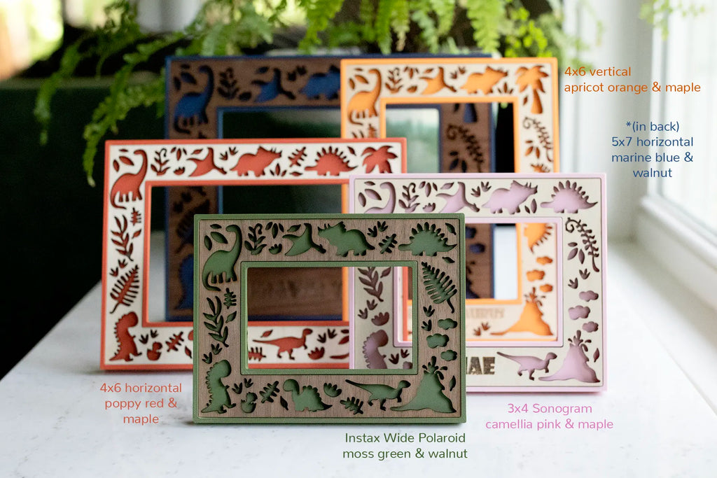 A collection of wooden picture frames for kids rooms with dinosaur laser-cut features in various colors, including orange, red, blue, green, and pink. The frames come in different sizes: 4x6, 5x7, 3x4, and Instax Wide sizes. Featuring prehistoric botanical and dinosaur designs.