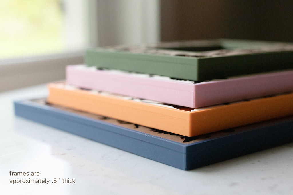 A stack of four colorful dinosaur picture frames in shades of blue, orange, pink, and green. The frames are approximately 0.5 inches thick. The frames are stacked on top of each other, with the blue frame at the bottom.