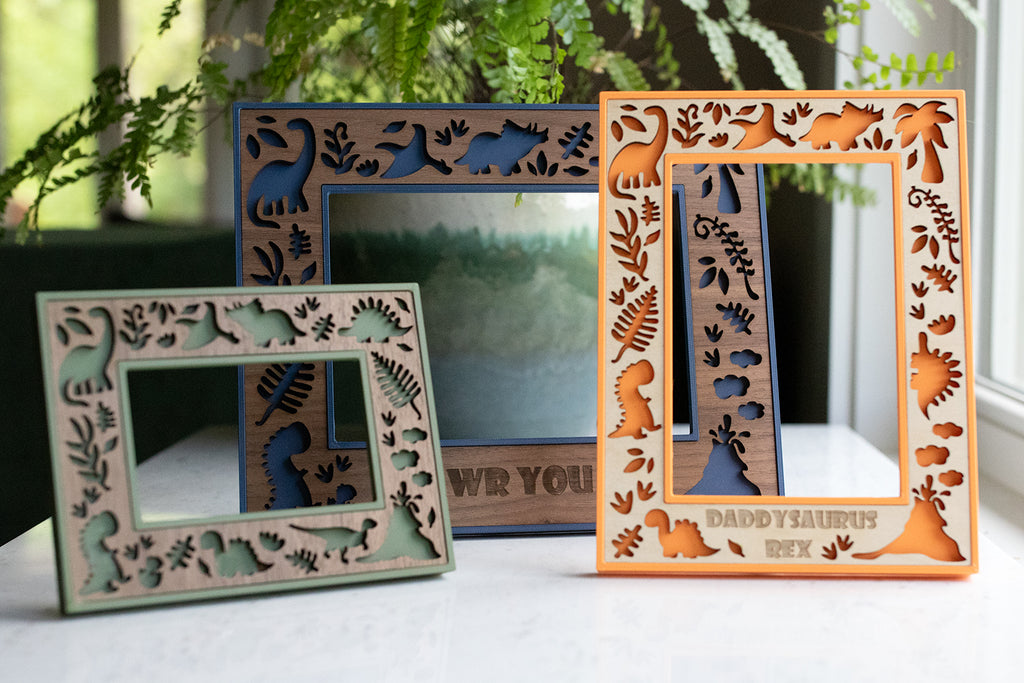 Three wooden picture frames with cute dinosaur cutouts. One frame is green, one is orange, and one is blue. The orange frame shows personalization with the words "Daddysaurus Rex". The frames are on a white surface with greenery in the background.