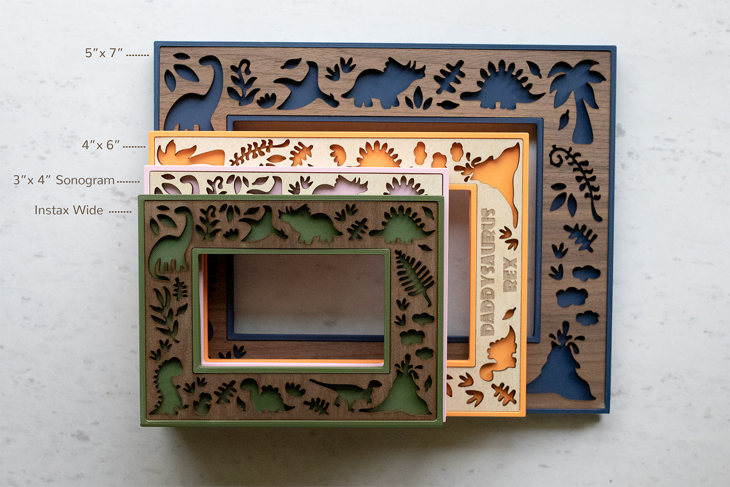 A stack of laser cut dinosaur picture frames in various sizes, each with a cute dinosaur theme. The largest frame is 5" x 7" with a blue border, while others include sizes 4" x 6", 3" x 4" Sonogram, and Instax Wide. The frames feature cut-out dinosaur and plant designs.