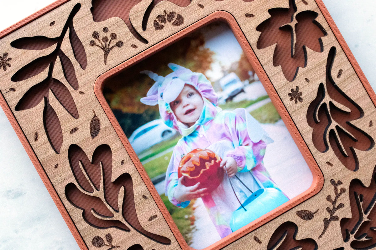 Close-up of the terracotta and cherry wood fall leaves magnetic frame, showing layered laser-cut autumn leaf cutouts and engraved acorn and berry details around an Instax Mini Halloween photo of a toddler in costume.