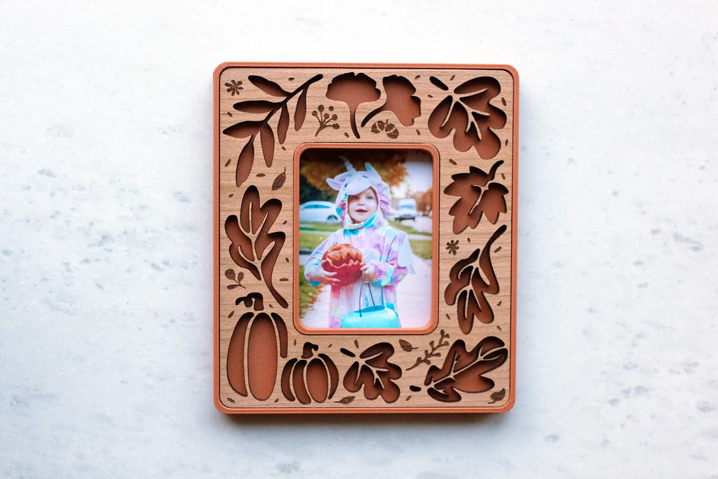 Terracotta and cherry wood fall leaves magnetic frame for Instax Mini photos, with laser-cut autumn leaf, ginkgo, and pumpkin cutouts, holding a Halloween photo of a toddler in costume with a jack-o’-lantern pail.