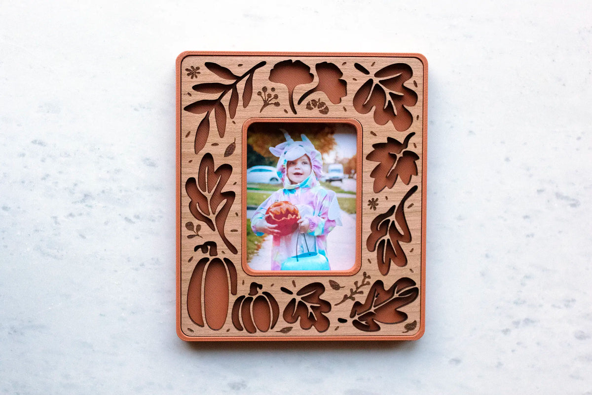 Terracotta and cherry wood fall leaves magnetic frame for Instax Mini photos, with laser-cut autumn leaf, ginkgo, and pumpkin cutouts, holding a Halloween photo of a toddler in costume with a jack-o’-lantern pail.
