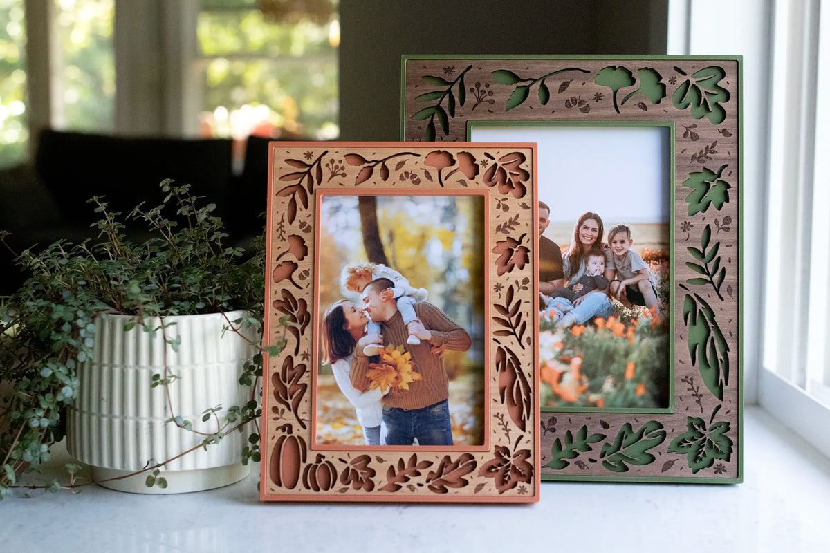 Two fall leaf picture frames on a bright windowsill — a 4×6 in terracotta with a cherry wood inlay in front and a 5×7 in moss green with a walnut inlay behind. Laser-cut with autumn leaves, pumpkins, and acorns, each holding a fall family portrait — a handmade way to display your seasonal autumn family photos as Thanksgiving and fall home decor.