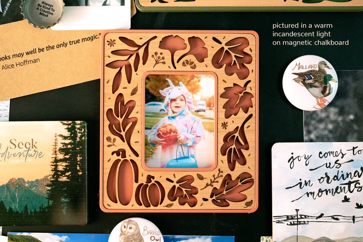 Terracotta and cherry wood fall leaves magnetic frame shown in warm incandescent light on a magnetic chalkboard among postcards and magnets, holding an Instax Mini Halloween photo of a toddler in costume.