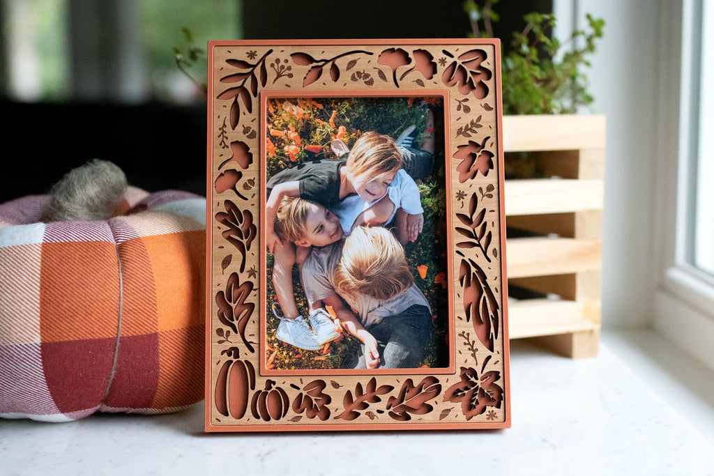 Terracotta laser-cut fall leaves and pumpkin picture frame holding a photo of three children lying in a flower field, styled with a small plaid handmade pumpkin and a plant.