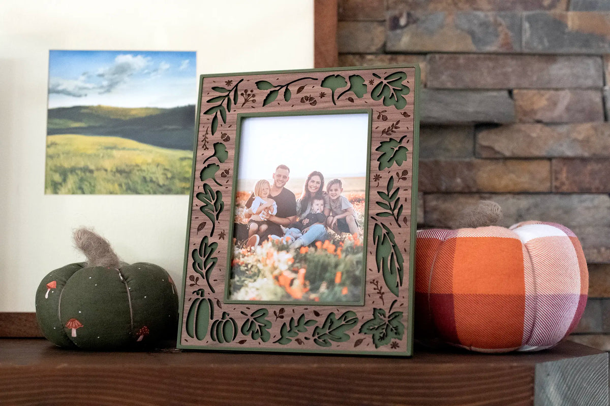 Moss green fall picture frame with a laser-cut walnut wood inlay, featuring autumn leaves, pumpkins, and acorns, displaying a fall family portrait of a family in a flower field. Styled on a mantel with fabric pumpkins — handmade fall and Thanksgiving home decor for your seasonal autumn family photos.