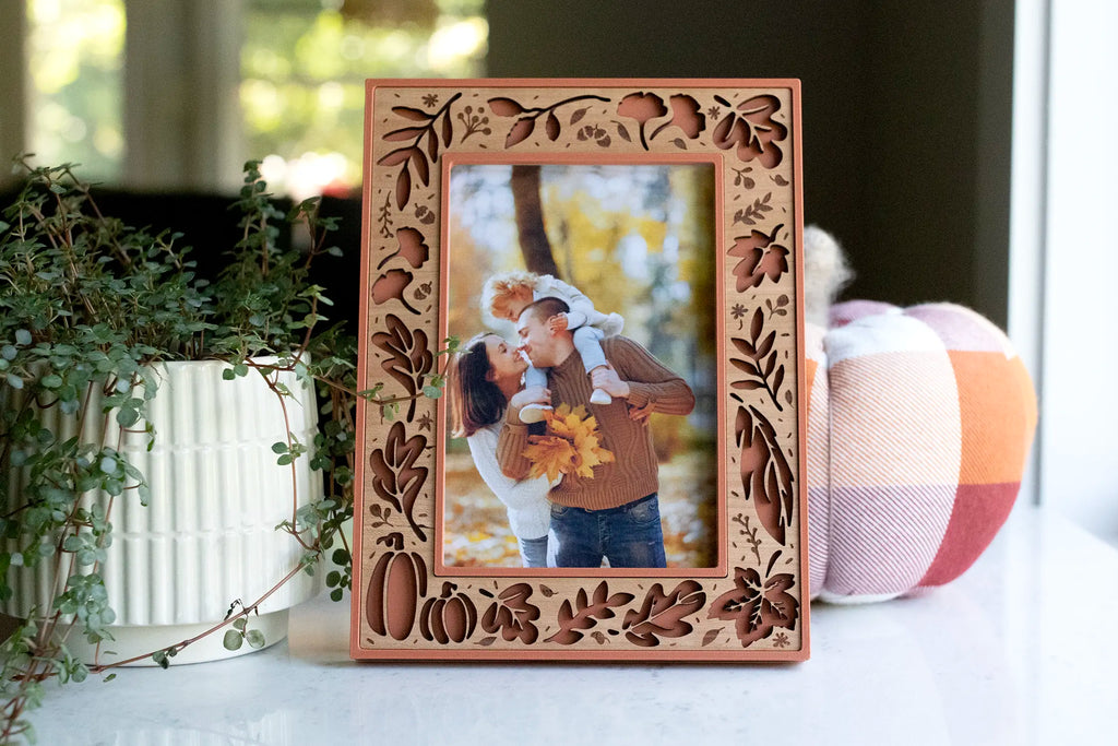 Terracotta fall picture frame with a laser-cut cherry wood inlay, featuring autumn leaves, branches, berries, and pumpkins — a personalized piece of fall home decor holding a family portrait in the autumn woods. Styled on a white counter with a trailing plant and plaid pumpkin as seasonal fall decor.