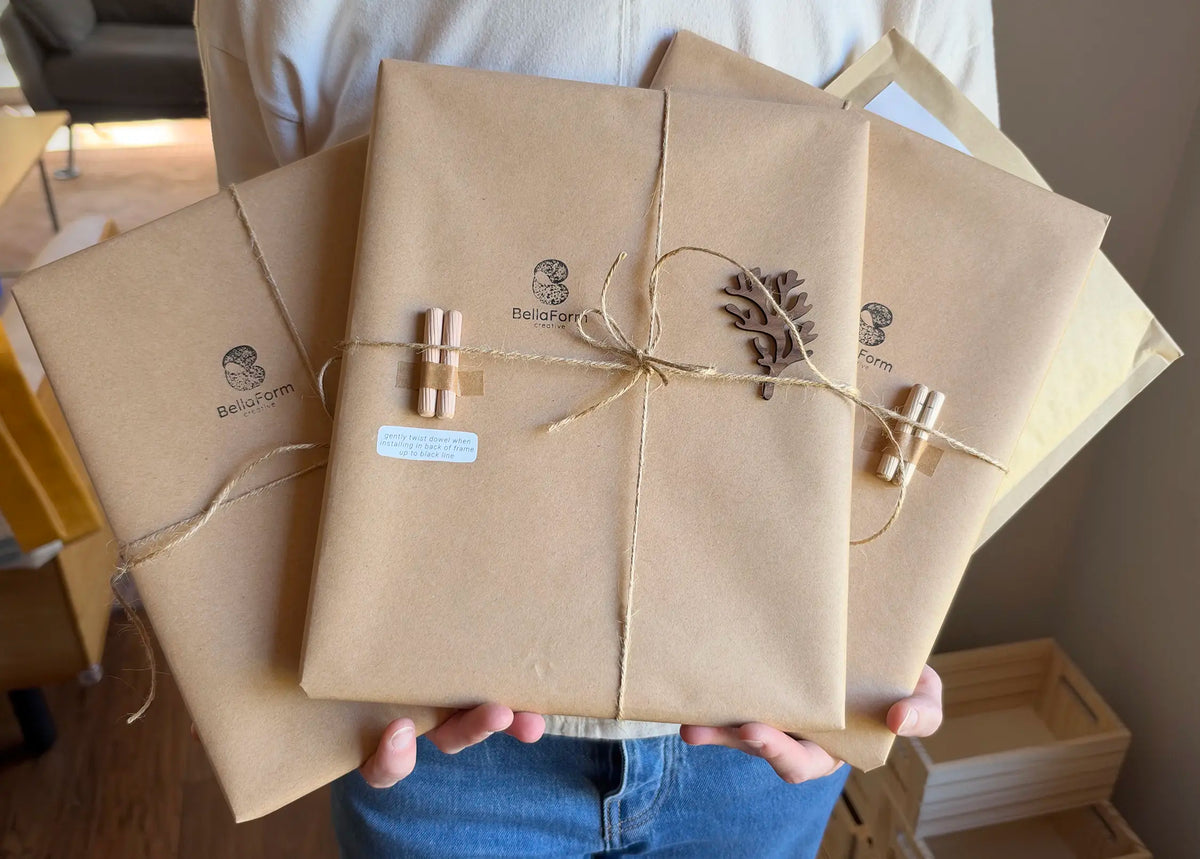 Three picture frame gifts wrapped in recycled kraft paper, each stamped with the BellaForm Creative logo and tied with natural jute twine, held out in front of a person in a white sweater. Small wooden dowels with a short instruction label are taped to the packages, and a laser-cut wooden coral-shaped accent is tied onto the center package.