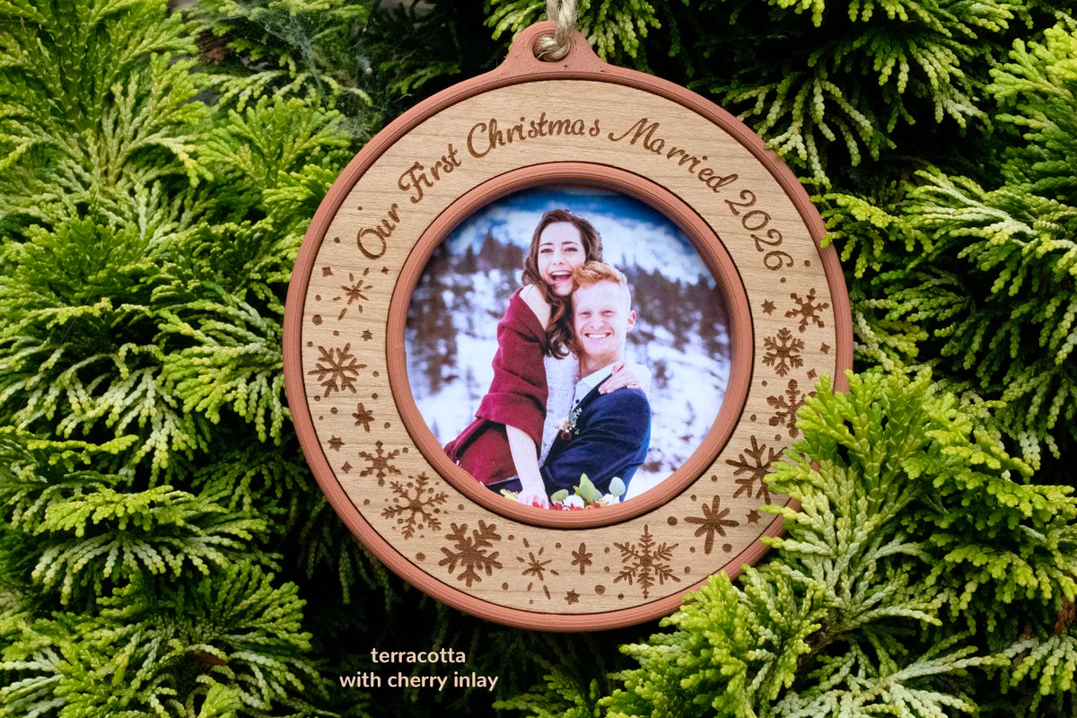 First Christmas married ornament in terracotta with a cherry inlay, laser-engraved "Our First Christmas Married 2026" with snowflakes, holding a wedding photo of a newlywed couple in the snowy mountains, hanging on a Christmas tree.