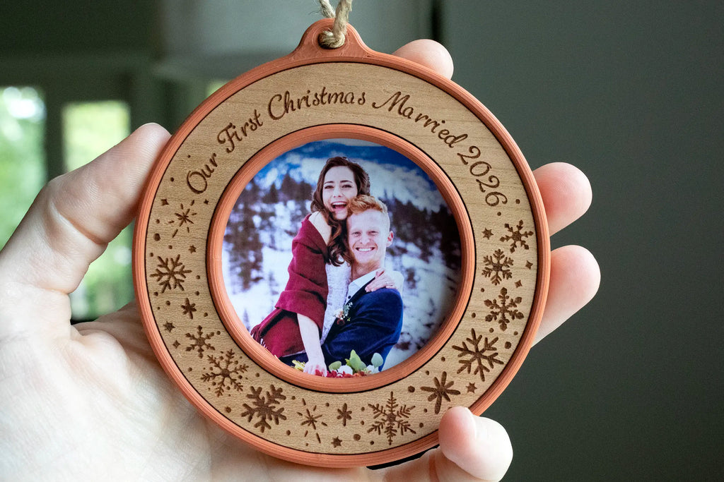 A 3.5 inch first Christmas married ornament in terracotta with a cherry wood inlay, laser-engraved "Our First Christmas Married 2026" with snowflakes. It's a mini picture frame holding a wedding photo of a newlywed couple in the snowy mountains and is being held to show scale.