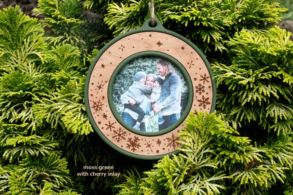 Christmas picture frame ornament in moss green with a cherry inlay, laser-engraved with snowflakes and falling snow and no text, holding a photo of a family of four hugging outdoors, hanging on evergreen branches.
