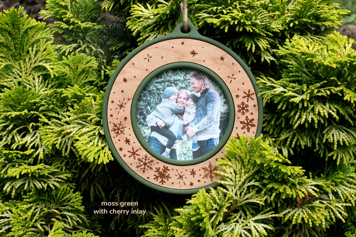 Christmas picture frame ornament in moss green with a cherry inlay, laser-engraved with snowflakes and falling snow and no text, holding a photo of a family of four hugging outdoors, hanging on evergreen branches.
