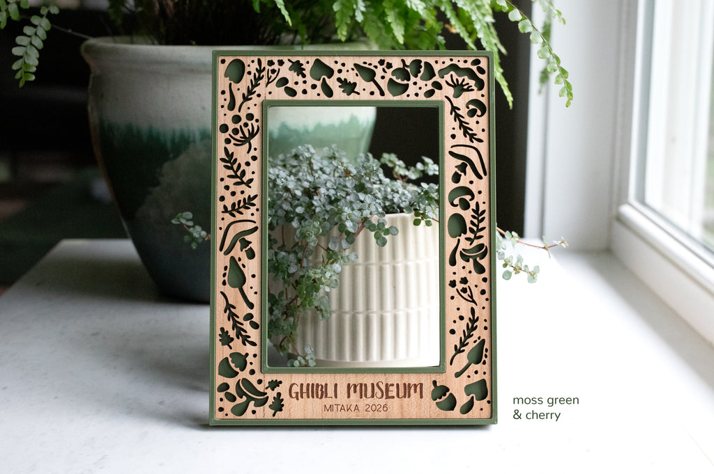 Dark green mushroom frame with engraving that reads "Ghibli Museum Mitaka 2026". 4x6 picture frame with custom engraving on wood. The frame has a delicate laser cut mushroom pattern, and there is greenery in the background.