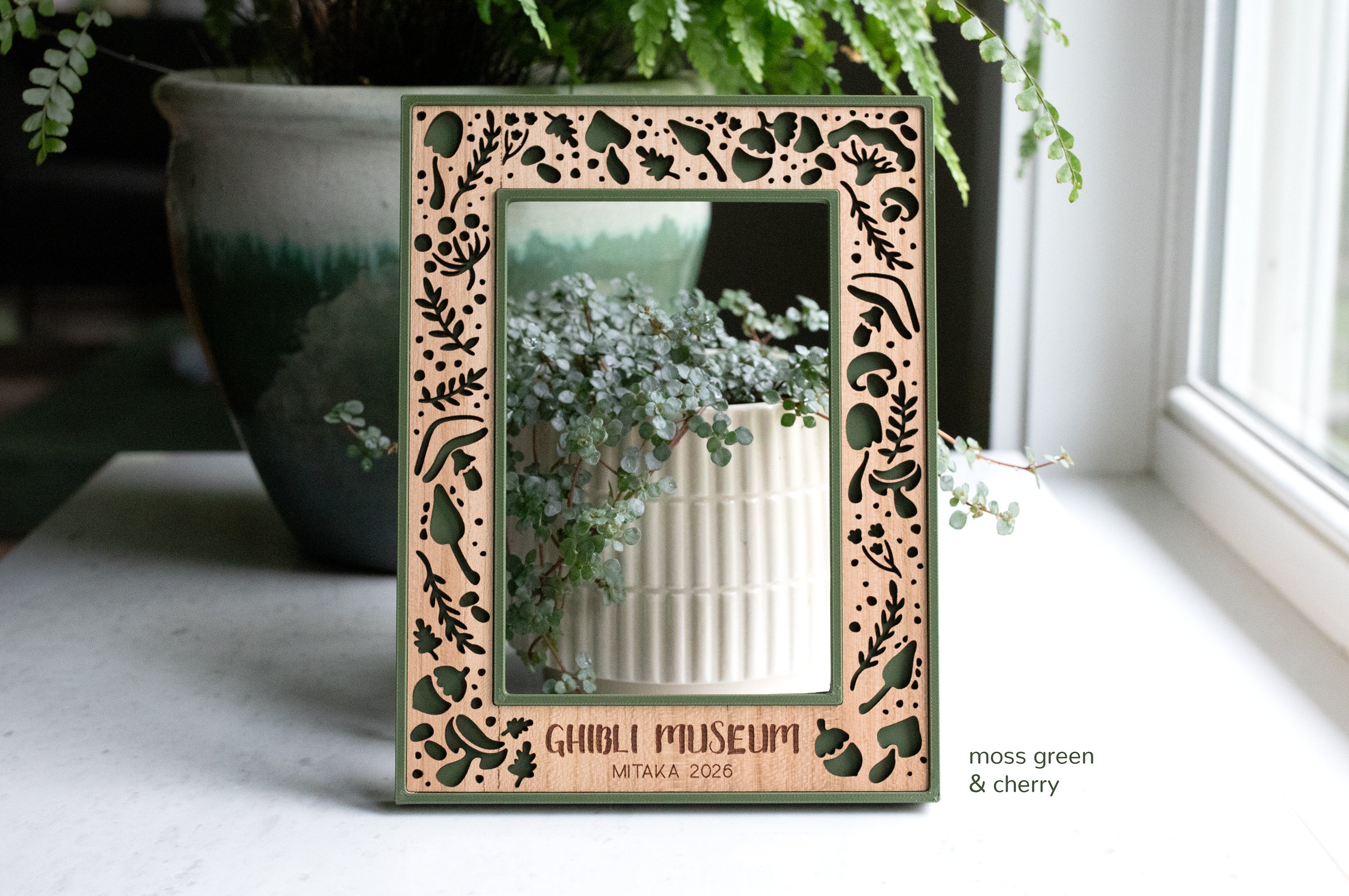 Dark green mushroom frame with engraving that reads "Ghibli Museum Mitaka 2026". 4x6 picture frame with custom engraving on wood. The frame has a delicate laser cut mushroom pattern, and there is greenery in the background.