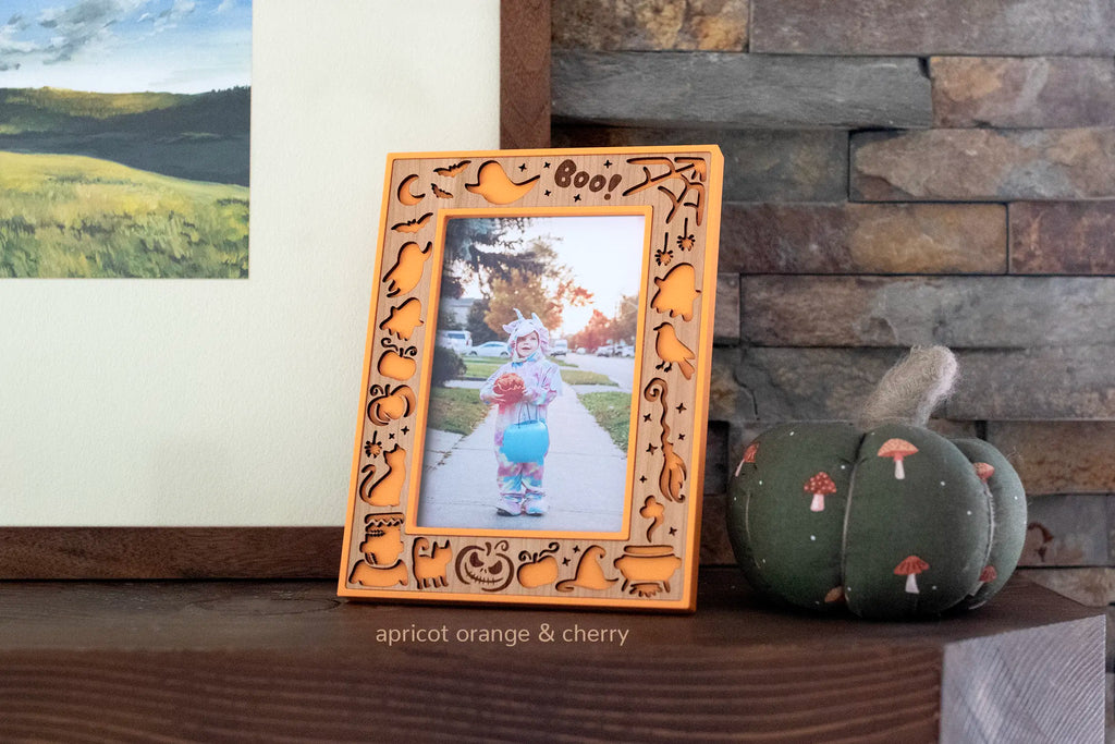 Apricot orange Halloween picture frame with a laser-cut cherry wood inlay, featuring ghosts, pumpkins, bats, a black cat, witch's hat, and cauldron around an engraved "Boo!", holding a photo of a child in a unicorn costume trick-or-treating. Displayed on a wooden mantel beside a green pumpkin.