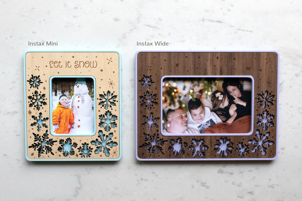 Two snowflake magnetic frames side by side on white marble — an Instax Mini (left) and an Instax Wide (right) — with laser-cut wood inlays featuring snowflakes, and blue and periwinkle frame housings, holding Christmas photos, one engraved "Let It Snow." A personalized Christmas keepsake and holiday stocking stuffer, offered in walnut, cherry, or maple and cool winter colors. Fits Instax instant film, or trim a regular photo to size.