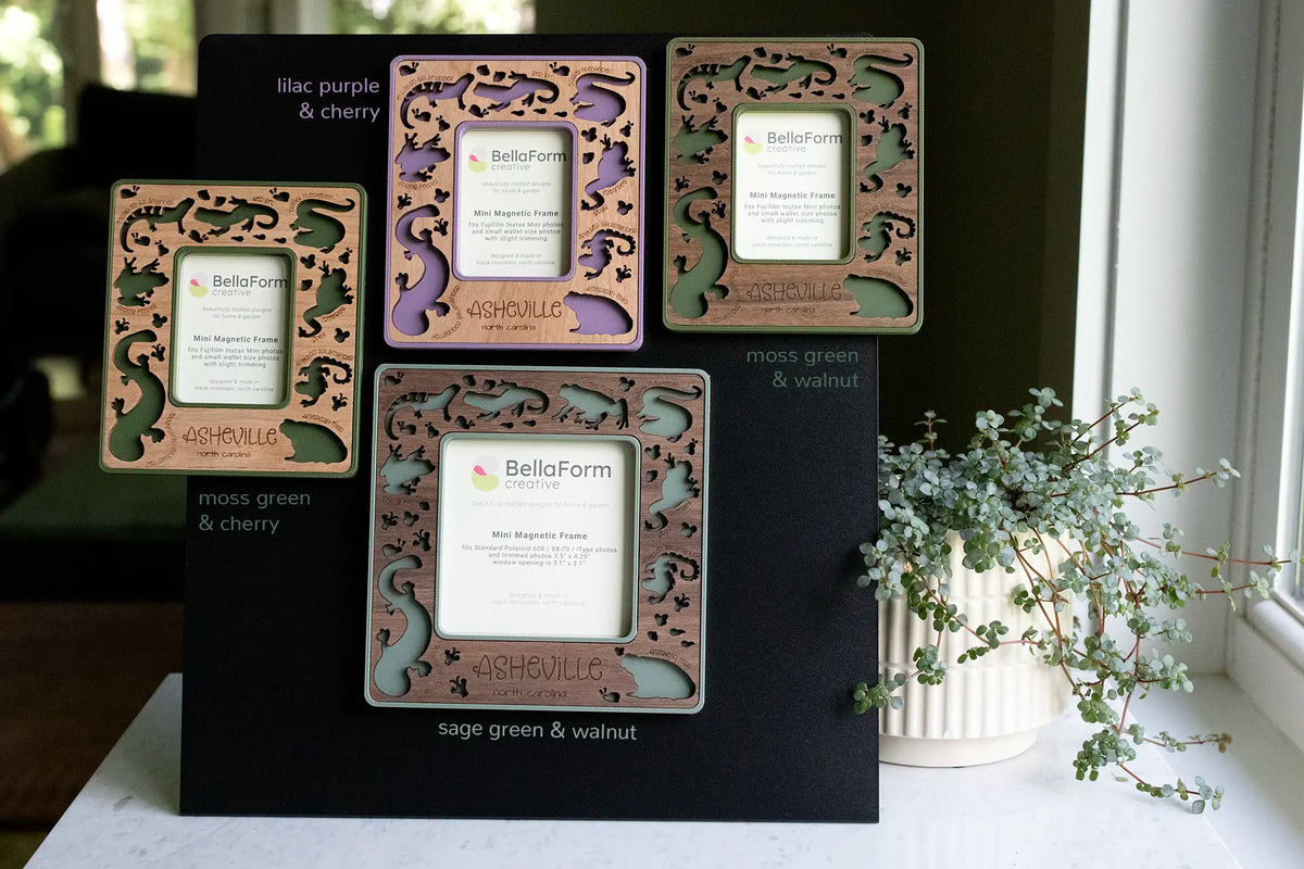 Four magnetic Appalachian amphibians frames in moss green and cherry, lilac purple and cherry, moss green and walnut, and sage green and walnut, with laser-cut salamander, frog, and toad cutouts and “Asheville, North Carolina” engraving, shown in Instax Mini and larger Polaroid sizes on a black magnetic board.