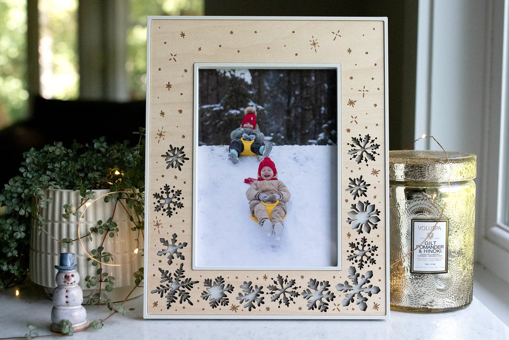 A large 5x7 holiday picture frame featuring delicate laser-cut snowflakes and engraved falling snow. The frame is linen white with a maple wood inlay. Surrounded by candles, lights, and winter holiday decor.