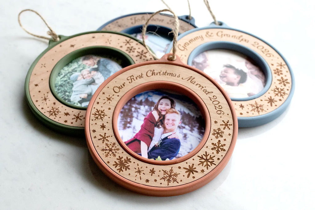 Stack of four personalized Christmas picture frame ornaments with jute twine hangers, led by a terracotta and cherry "Our First Christmas Married 2026" ornament with a newlywed couple's photo. Behind it are moss green, slate blue "Grammy & Grandpa 2026," and marine blue family photo ornaments, all laser-engraved with snowflakes and falling snow.