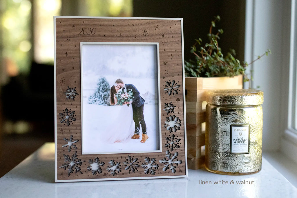 Laser cut snowflake picture frame with personalized "2026" engraved in the top left corner. Frame is linen white with a walnut wood inlay, featuring delicate snowflakes and displaying a photo of a couple at their winter wedding.