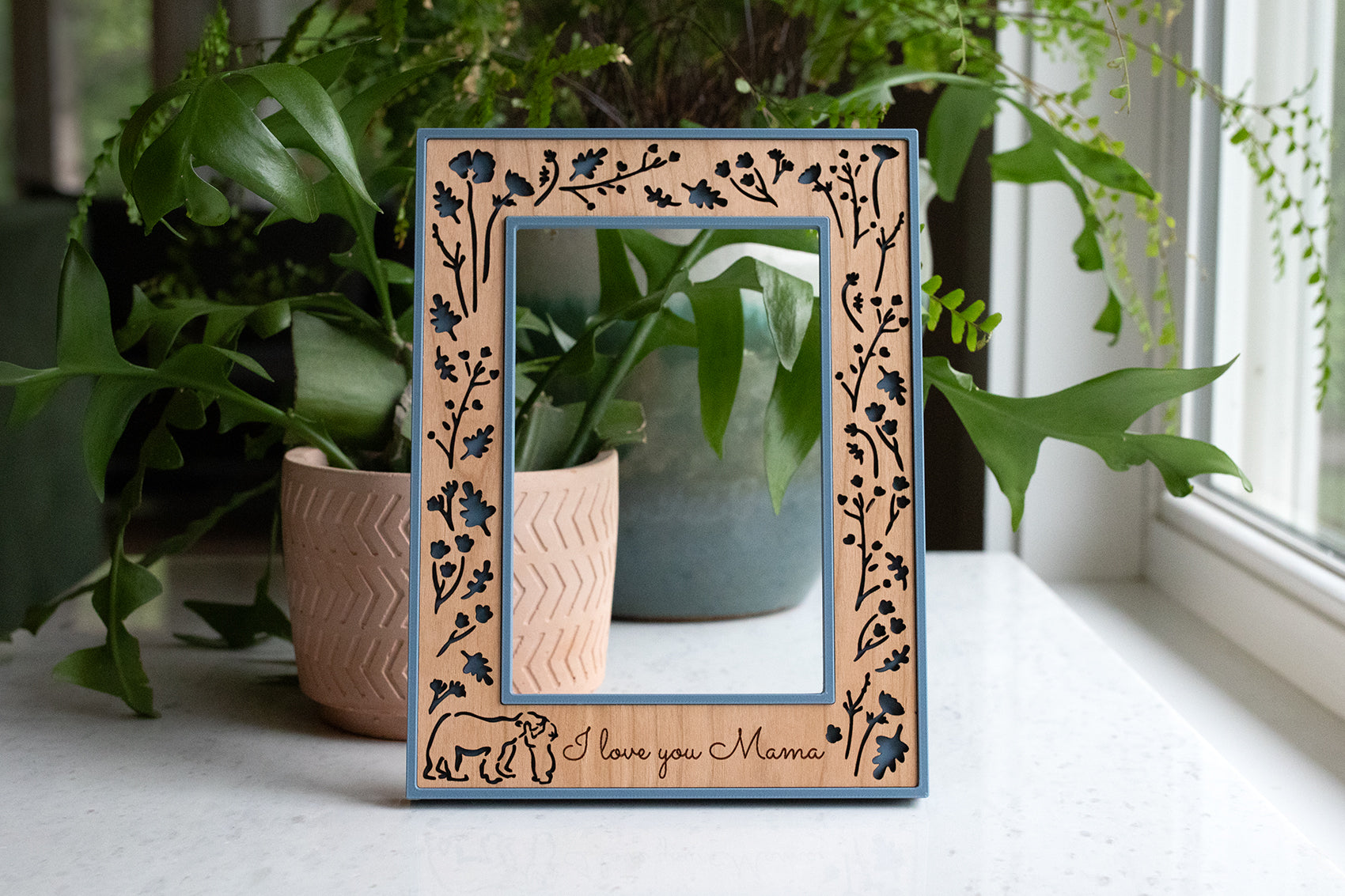 May include: Wooden picture frame with a light blue border and floral cutouts. The frame has a bear and the text "I love you Mama" engraved on the bottom. The frame is displayed in front of green plants.