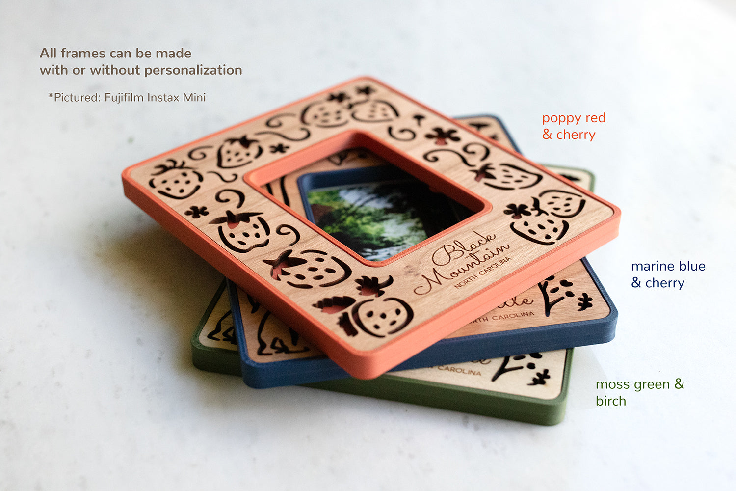 Three Instax Mini magnetic Polaroid photo frames stacked on top of each other. The top left corner of the image displays the words: "All frames can be made with or without personalization". The frames have a moss green border, a navy blue border, and a neutral red color. The wooden inlays are cherry and birch.