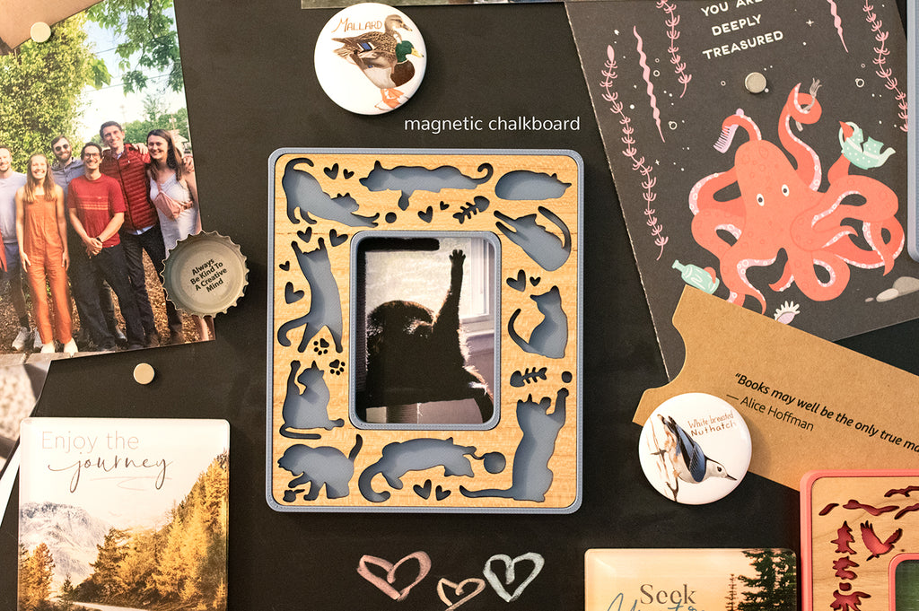 A tiny blue magnetic cat frame for cat lovers hanging on a magnetic chalkboard. The frame is surrounded by postcards and photographs. The frame holds Instax Mini size photographs or photos trimmed to size.
