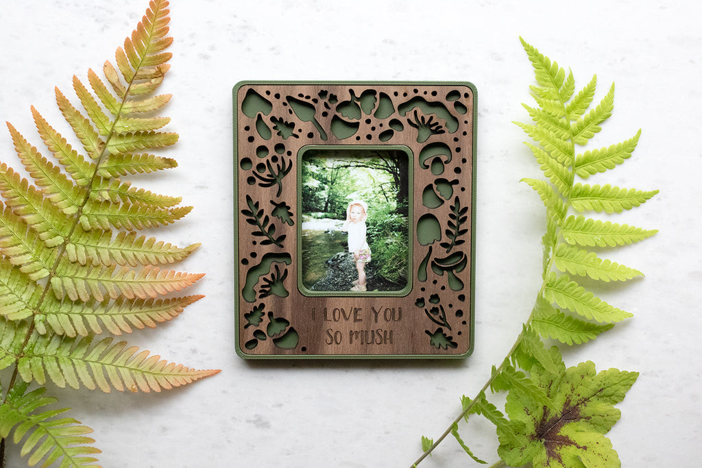 A whimsical magnetic frame with a mushroom and acorns design. The mini frame is laser cut from walnut and includes a moss green color border. The frame holds an Instax Mini photograph and is displaying a picture of a little girl. Personalized engraving reads "I love you so mush". There are ferns and greenery around the frame.