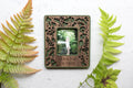 A whimsical magnetic frame with a mushroom and acorns design. The mini frame is laser cut from walnut and includes a moss green color border. The frame holds an Instax Mini photograph and is displaying a picture of a little girl. Personalized engraving reads "I love you so mush". There are ferns and greenery around the frame.