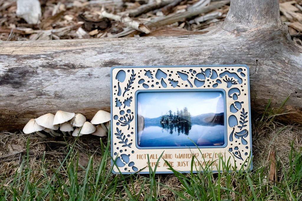 A mushroom magnetic instax wide frame in blue with a wooden inlay. The frame is propped against a log in the grass next to a patch of wild mushrooms. The frame reads "not all who wander are lost, some are just foraging." A great gift for mushroom lovers.