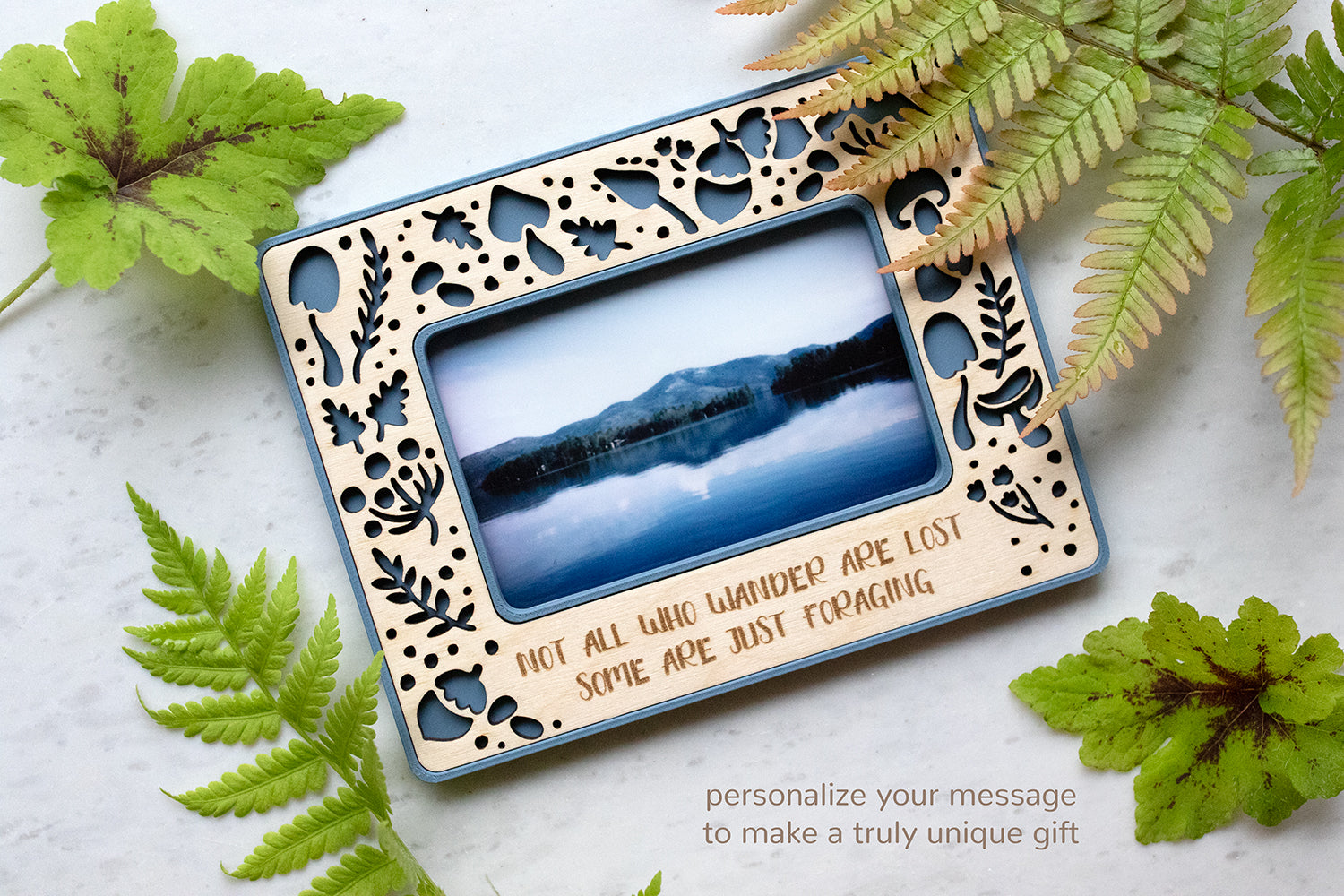 A blue mushroom frame design with a birch wood inlay surrounded by greenery. The personalized message at the bottom of the frame reads "Not all who wander are lost, some are just foraging." The Instax Wide frame has a playful, whimsical aesthetic.
