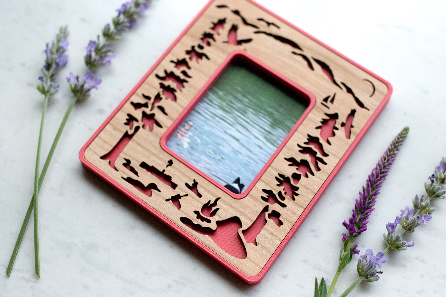 Instax Mini Polaroid photo frame in pink with a laser cut cherry wood inlay, featuring Lake George loons, osprey, and mountains. The frame is surrounded by wildflowers and lying on a white surface. Mini frame displays a photograph of a woman and a dog swimming in Lake George.