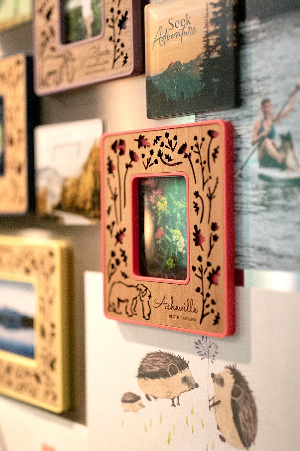 A close-up detail view of an Instax Mini frame on the front of a refrigerator. The frame has an intricate laser cut design, displaying woodland flowers and baby bear hugging mama bear. The words "Asheville North Carolina" are engraved in the wooden inlay, and the frame border is a bright pink color.