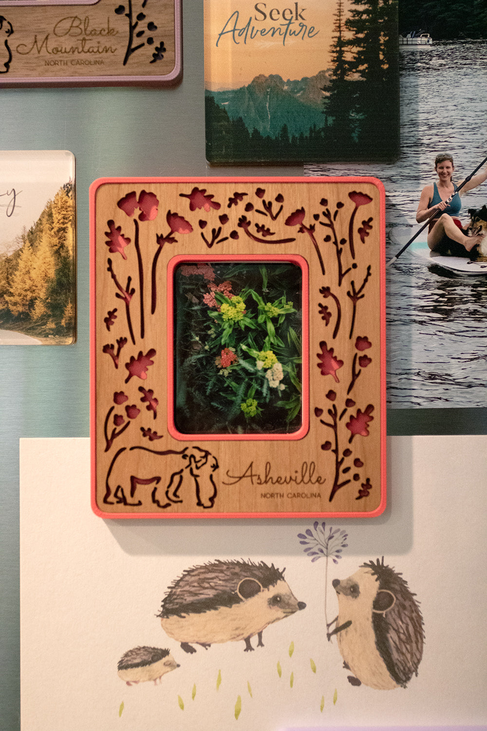 A close-up detail view of an Instax Mini frame on the front of a refrigerator. The frame has an intricate laser cut design, displaying woodland flowers and baby bear hugging mama bear. The words "Asheville North Carolina" are engraved in the wooden inlay, and the frame border is a bright pink color.