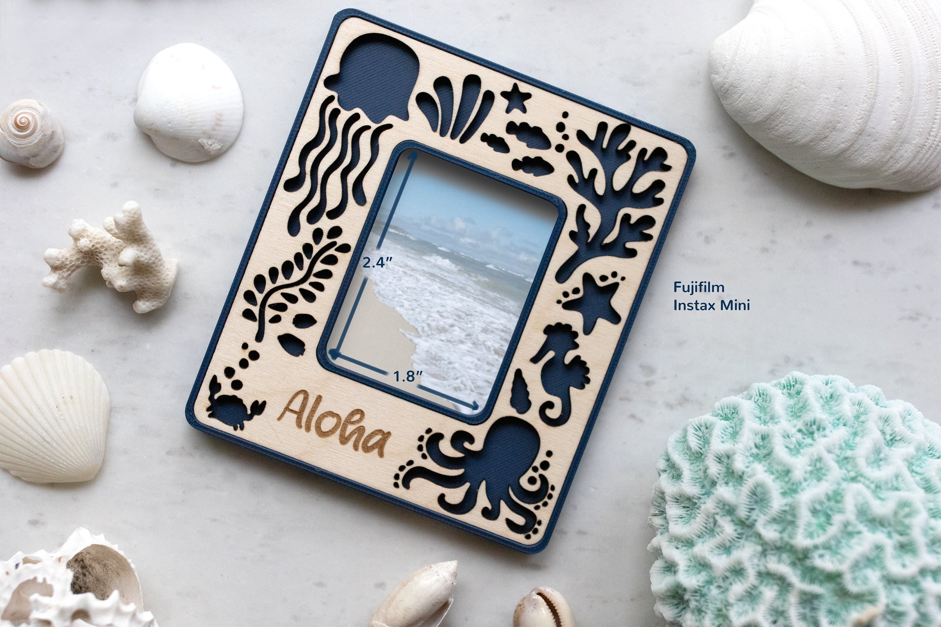 Instax mini beach frame that has a personalized "Aloha" message and cute sea creatures. The frame opening is 2.4" x 1.8" and displays a photograph of the beach and ocean. The frame is surrounded by seashells and a blue coral candle.
