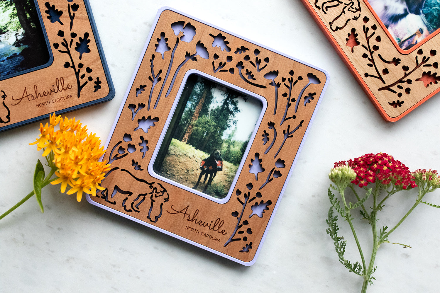 An assortment of magnetic Polaroid photo frames with laser cut woodland flowers and baby bear hugging mama bear. The frames are customizable with a variety of colors and wood types. The frames are lying amongst brightly colored flowers.
