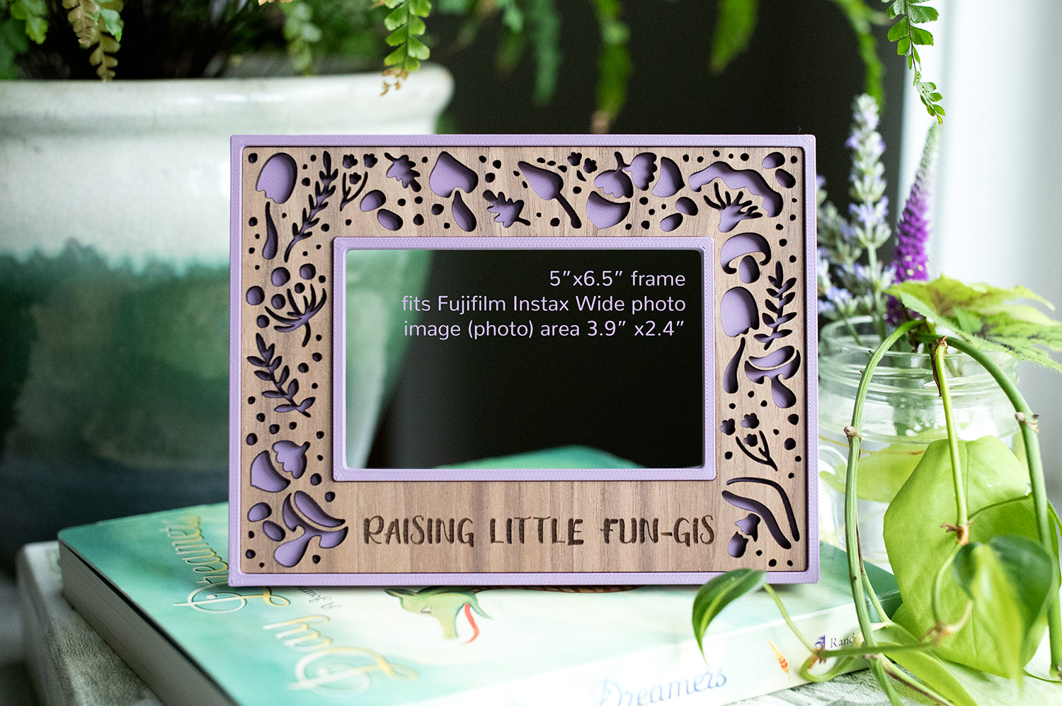 An Instax Wide polaroid frame that fits instax wide photos. The frame is purple with a laser cut walnut inlay featuring whimsical mushrooms and acorns. The personalized message on the frame reads: "raising little fun-gis". There is greenery around the frame.