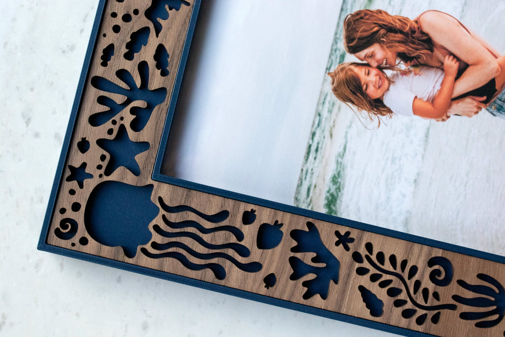 Close-up of a walnut frame corner with navy blue edging, showing laser-cut jellyfish, starfish, coral, and seashell cutouts.