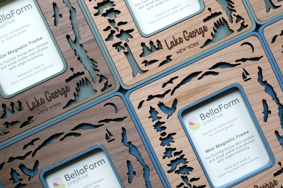Lake George magnetic frames with wooden inlays. Custom Lake George keepsakes for Adirondack lovers. Frames feature the silhouette of Lake George NY, with Adirondack mountains in the distance, loons on the lake, osprey flying in the sky, evergreens on islands in the lake, the famous Lac Du Saint Sacrement tour boat, and a pair of kayakers. Pictured in various shades of blue and green with walnut and cherry wood inlays.