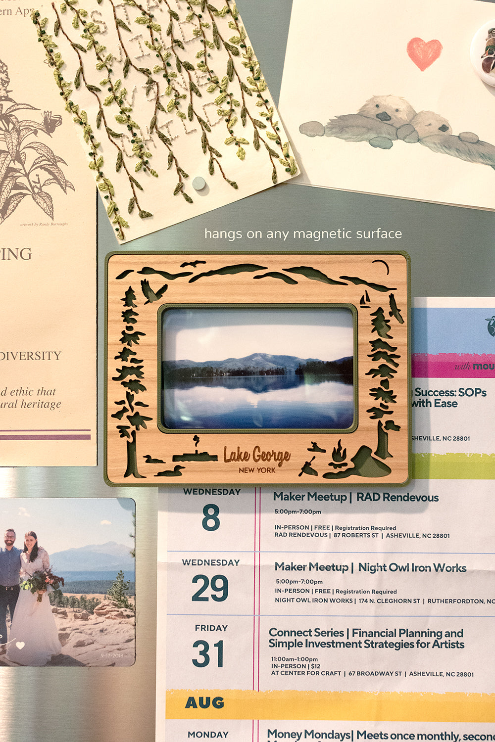 Lake George themed Instax Wide frame with magnets on a stainless steel refrigerator. The frame has an intricate, laser cut wooden inlay including imagery from Lake George New York - trees, osprey, birds, boats, and other lake decor elements. The frame is surrounded by cute postcards and photographs. The words "hangs on any magnetic surface" are visible.