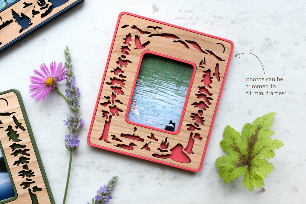A magnetic Instax Mini Frame with laser cut wooden inlay, featuring Lake George New York. The magnetic frame is surrounded by flowers and displaying a photo of a dog and a woman swimming in the lake.