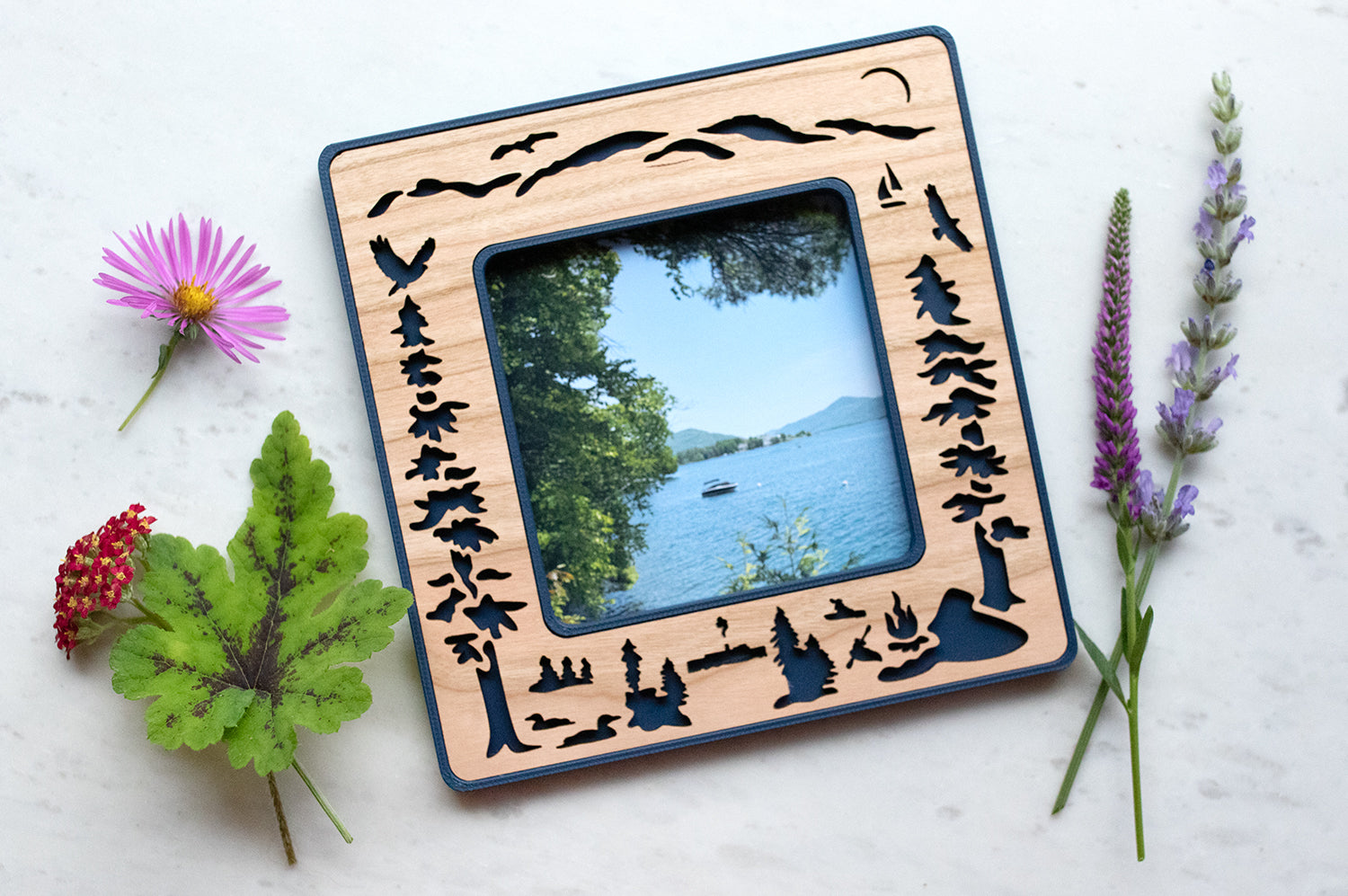 A navy blue and cherry wood Polaroid frame in Standard Polaroid photo size featuring a laser cut design of Lake George New York. The design includes evergreen trees, adirondack mountains in the background, and loons, kayakers, and a campfire in the foreground. The Polaroid frame fits standard 600 / SX-70 / iType photos.