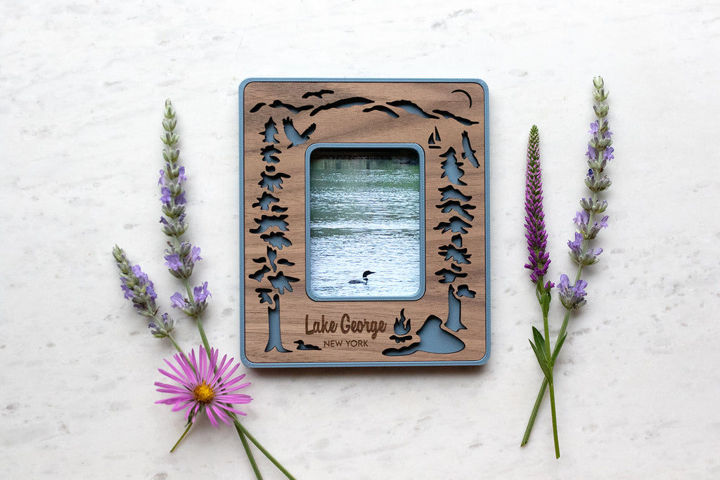 Lake George New York magnetic polaroid frame in Instax Mini frame size. The frame features laser cut Adirondack mountain, evergreen trees, loons, osprey, and kayakers on Lake George. The mini frame is lying next to wildflowers and displays a photo of a loon swimming on the lake.